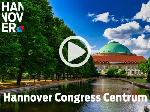 Hannover Congress Centrum mit grüner Kuppel, rotem Dach und spiegelndem Wasserbecken, umgeben von Bäumen unter blauem Himmel.